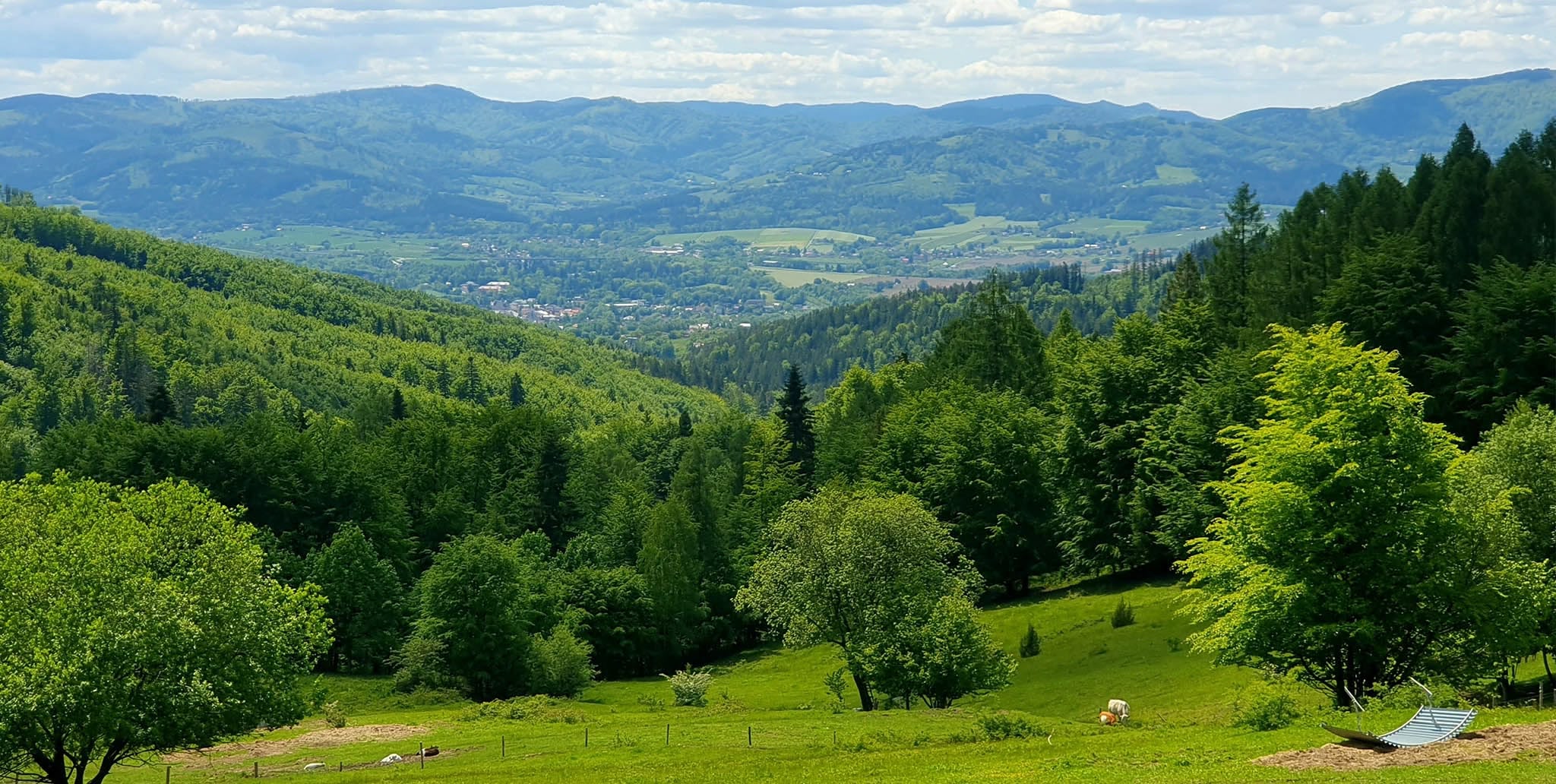 Zdjęcie przedstawia krajobraz górski pełen zieleni. W oddali widać pomniejsze zabudowania, powietrze jest przejrzyste, niebo błękitne, jasne i delikatnie zachmurzone.