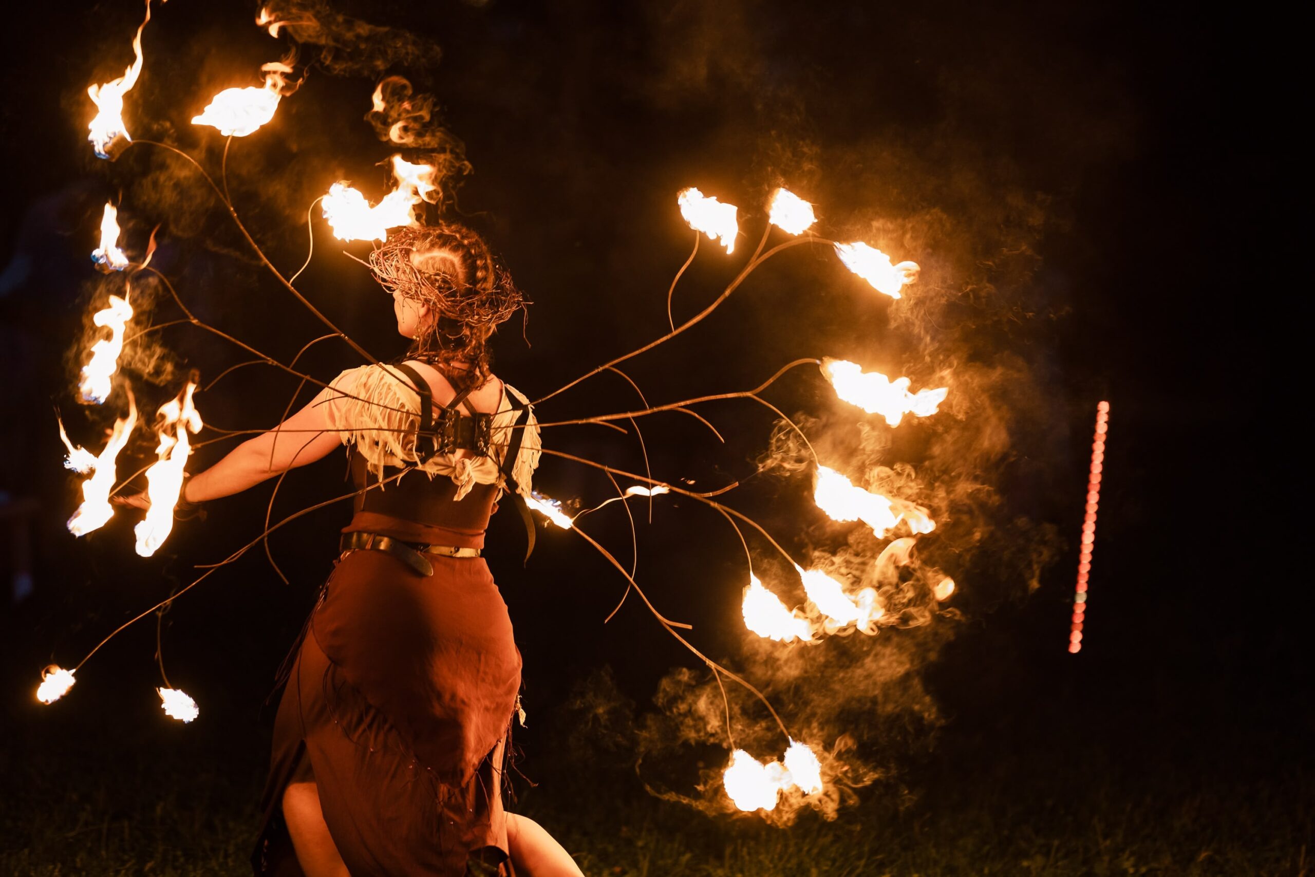 Fotografia przedstawia dziewczynę w wianku na głowie z przytwierdzonymi do pleców płonącymi patykami ułożonymi w skrzydła.